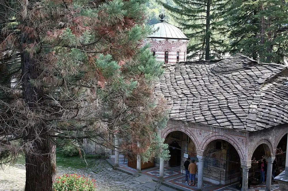 Wooden galleries and stone wings of Troyan Monastery around a green courtyard with tall trees
