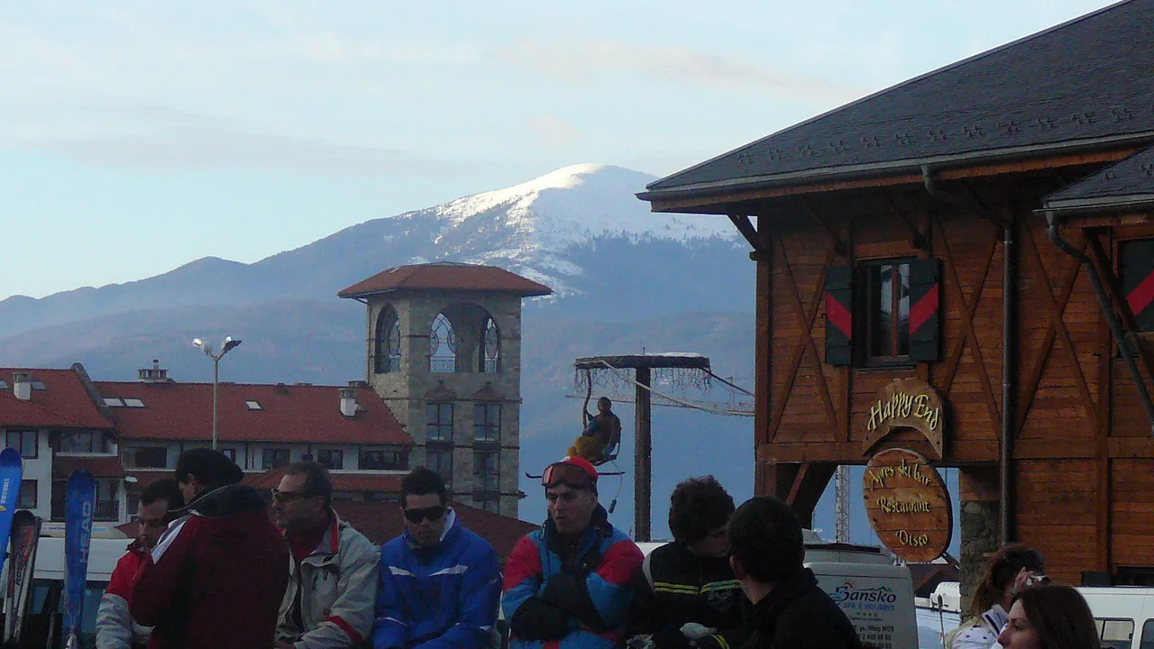 Skiers in jackets and goggles gathered outside the Happy End après-ski bar at the foot of the Bansko gondola, a snow-capped summit beyond the rooftops