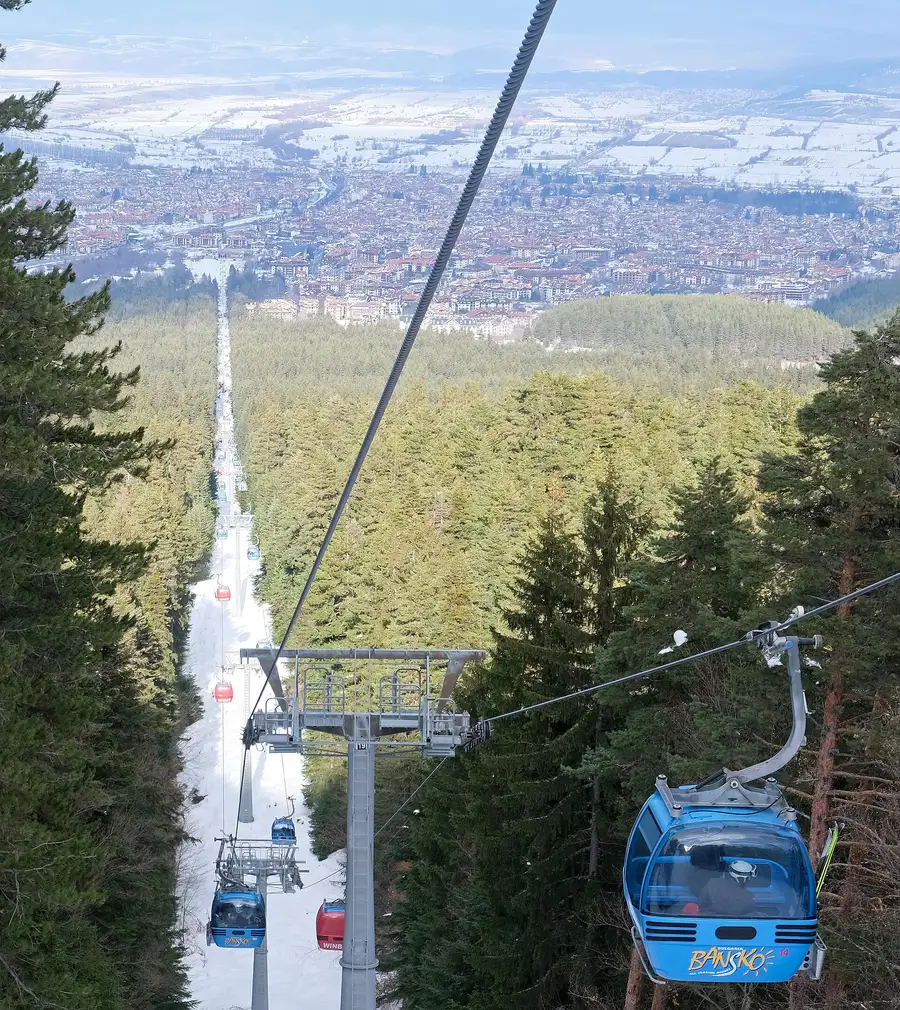 A blue gondola cabin descending over pine forest toward the town of Bansko spread across the valley floor below