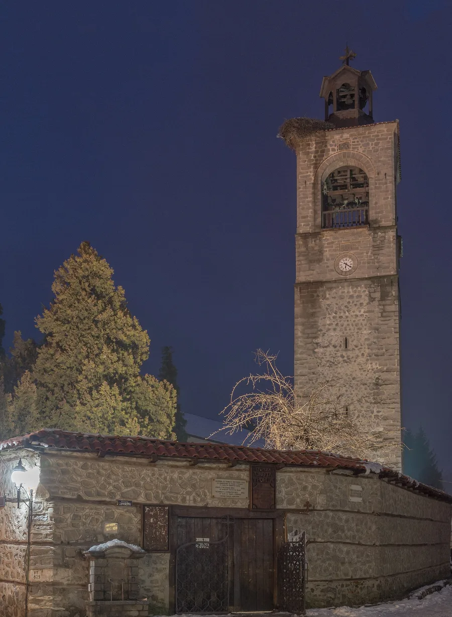 The stone bell tower of Holy Trinity church in Bansko lit up at night above the old church wall and its wooden gate, a little snow on the ground