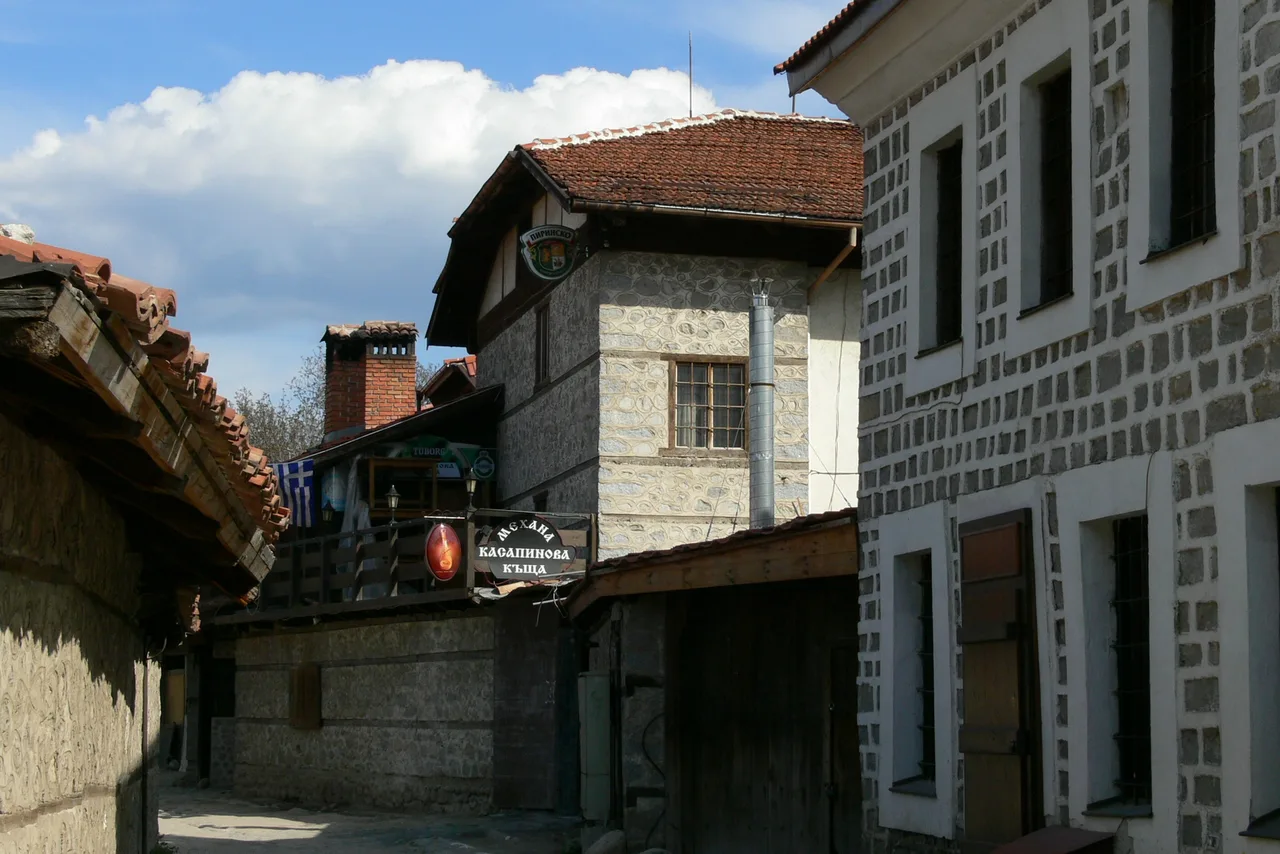 A narrow lane in Bansko Old Town between stone houses, with the hanging sign of Mehana Kasapinova Kashta and a Pirinsko beer sign above a wooden balcony