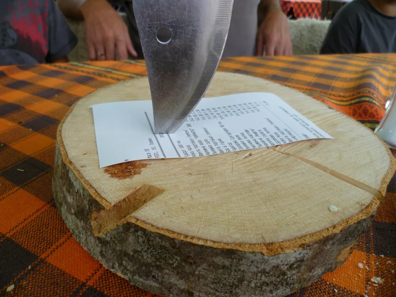 A printed restaurant bill pinned to a thick slice of log by the tip of a large knife, on a woven orange tablecloth