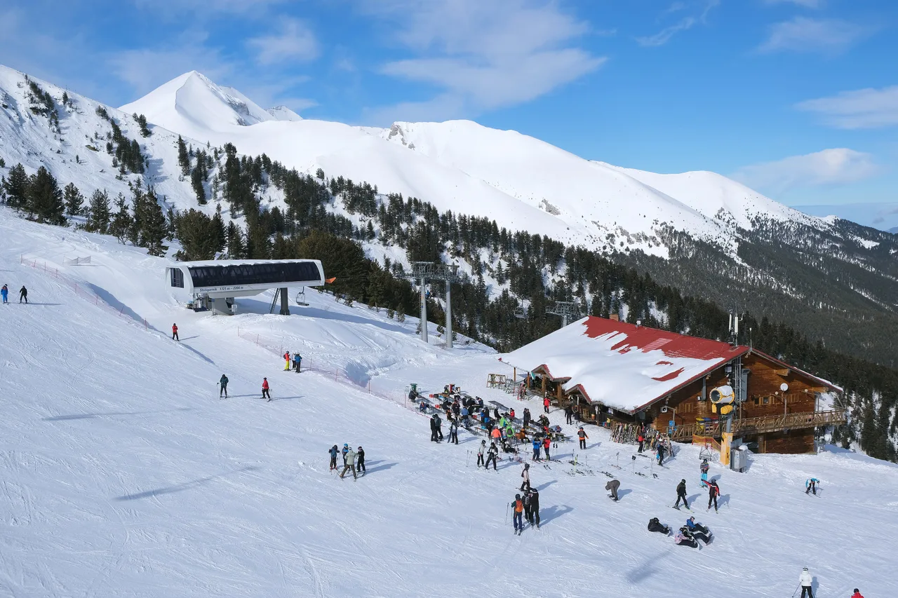 A wooden mountain hut with a snow-covered red roof beside the Shiligarnik chairlift station at Bansko, skiers resting at benches outside