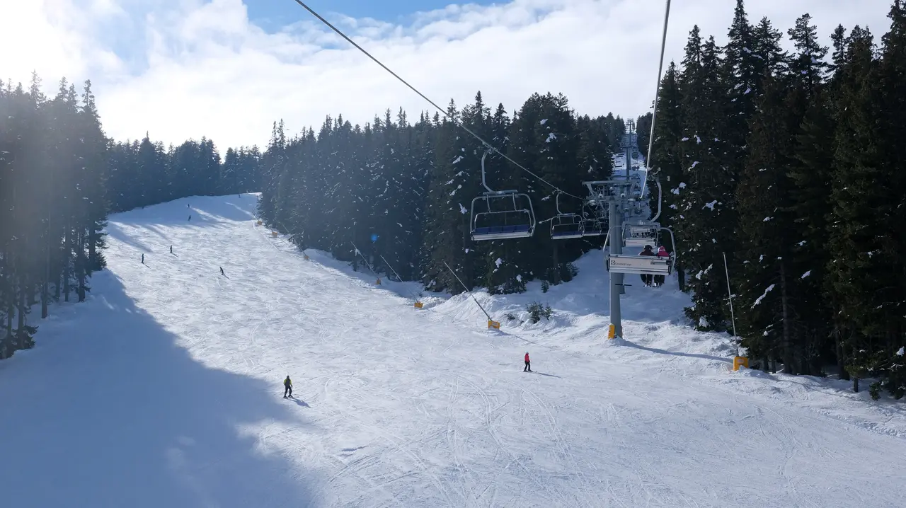 Skiers on a sunny groomed piste at Bansko with a chairlift running up the tree-lined slope