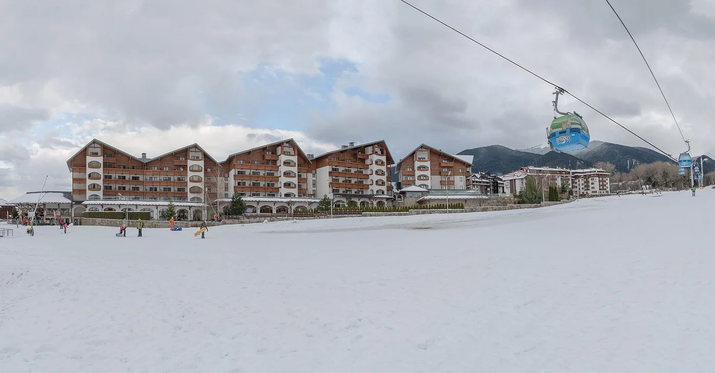 The town of Bansko under snow in winter, red-roofed houses spread across the valley with the white peaks of the Pirin mountains rising steeply behind