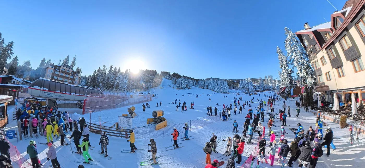 Wide groomed ski pistes at Borovets curving down through snow-dusted pine forest, with skiers on the runs and mountains in the distance