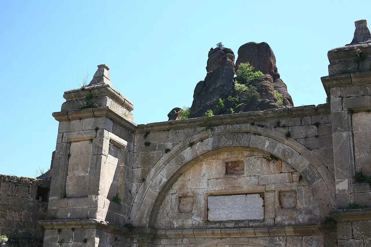 The ornate carved stone gateway of Belogradchik fortress with a rosette-decorated arch, red rock pinnacles rising behind the wall