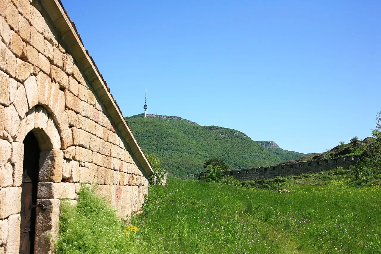 The stone ramparts of Belogradchik fortress running along a green ridge toward a wooded hill with a TV tower on top
