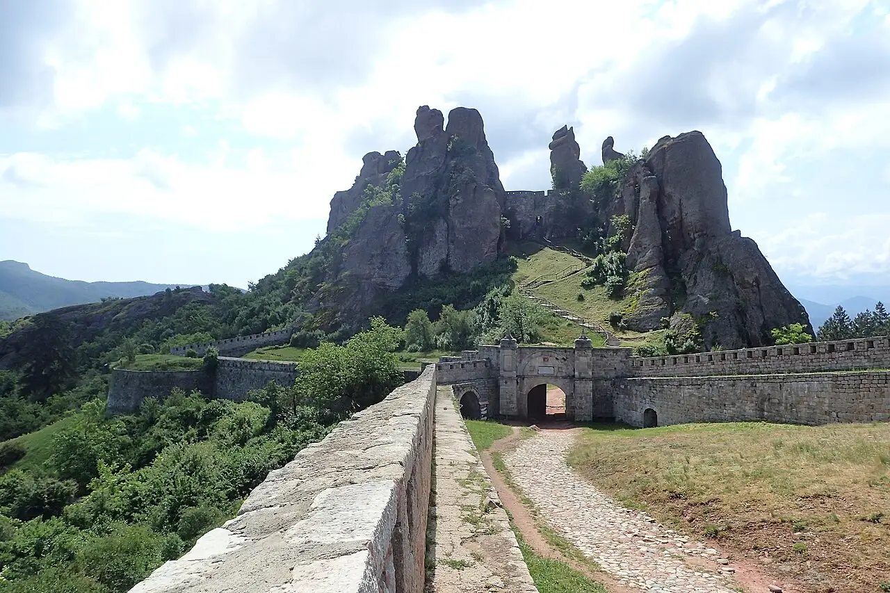 A stone wall walkway leading to the fortress gate, with a staircase climbing the grassy slope toward the towering rock pinnacles behind
