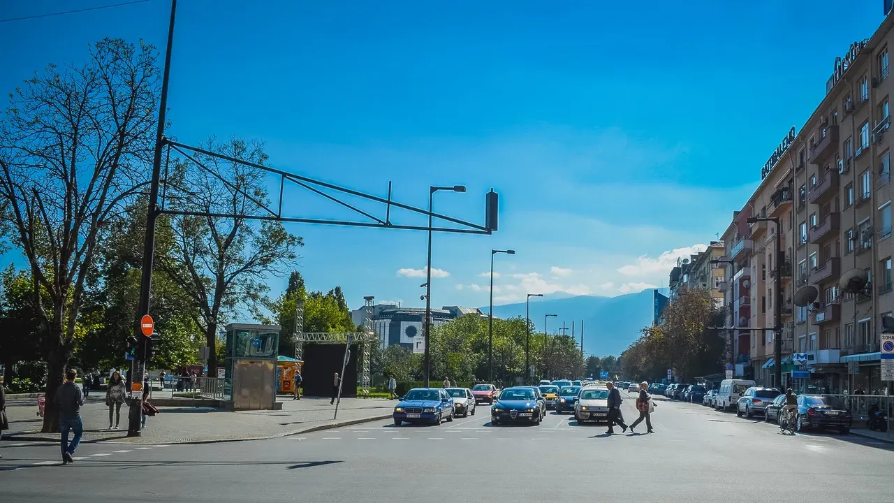 A busy central Sofia street with cars and pedestrians crossing under a blue sky, mountains visible in the distance
