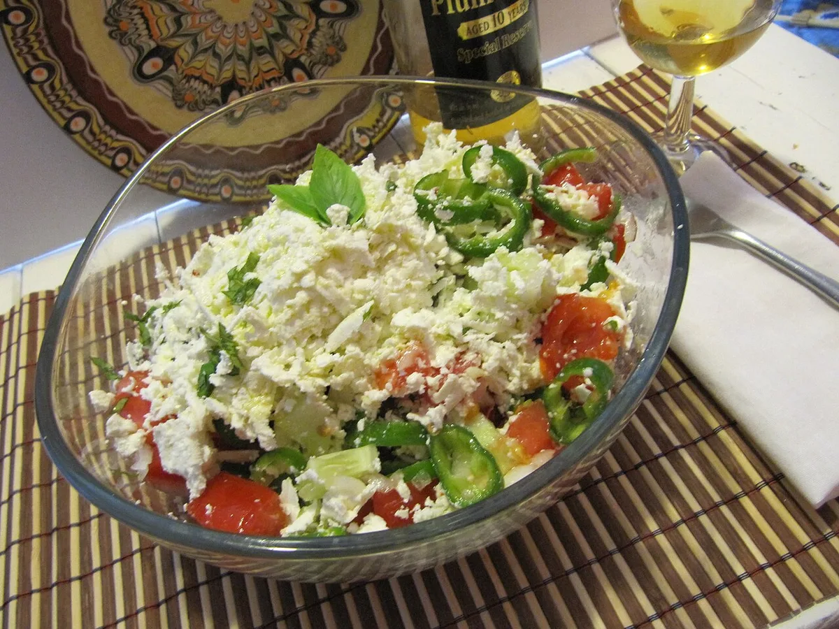 A shopska salad topped with grated white cheese beside a small glass of plum rakia on a Bulgarian table