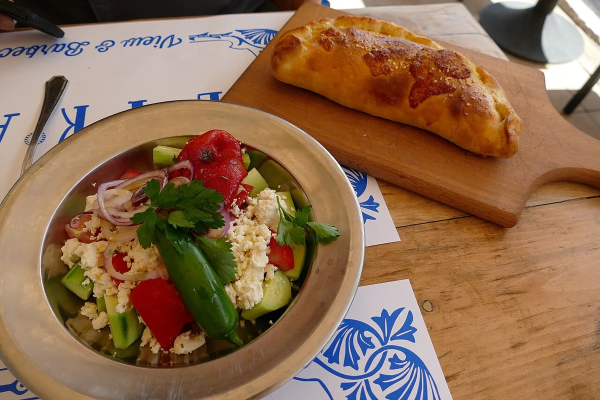 A shopska salad topped with grated white cheese and a green chili in a metal bowl, beside a golden baked bread on a board on a Sofia restaurant table