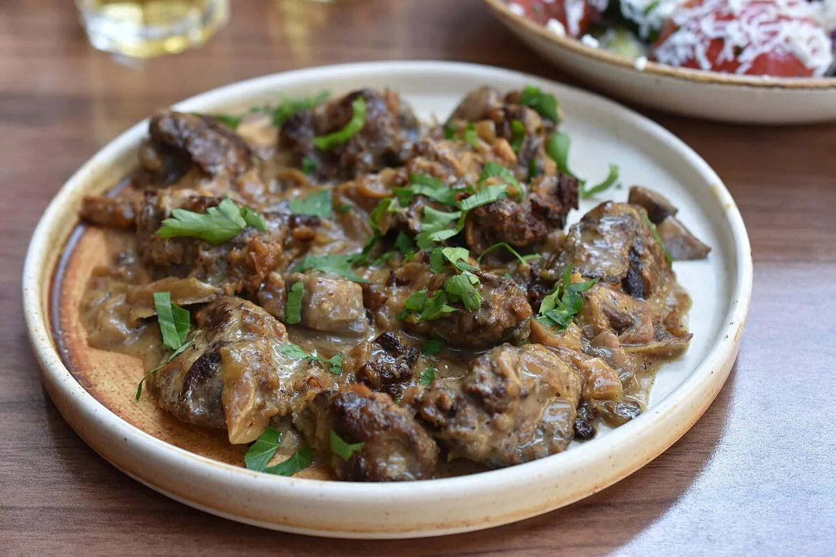 A plate of drob, a Bulgarian dish of diced offal in a creamy onion sauce topped with fresh parsley, with a salad and a glass of white behind