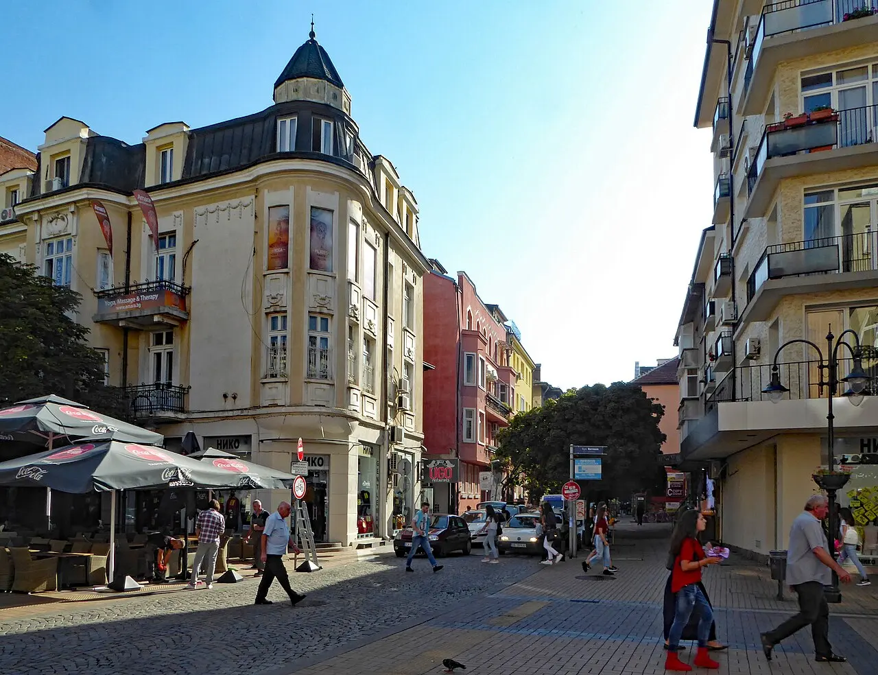 A pedestrian shopping street in central Sofia in the afternoon sun, with cafe umbrellas, an ornate corner building with a turret, and people walking
