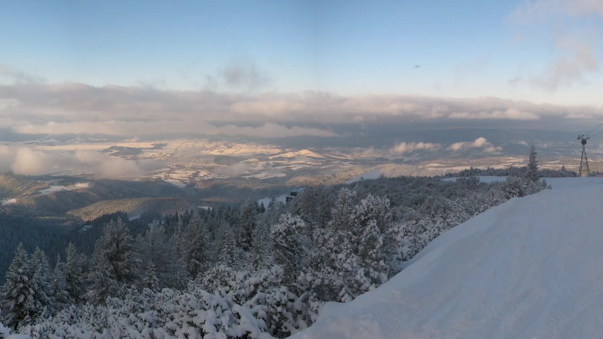 Snow-covered piste and gondola cable line above Borovets, with the forested Rila valley and distant hills spreading out below at dawn