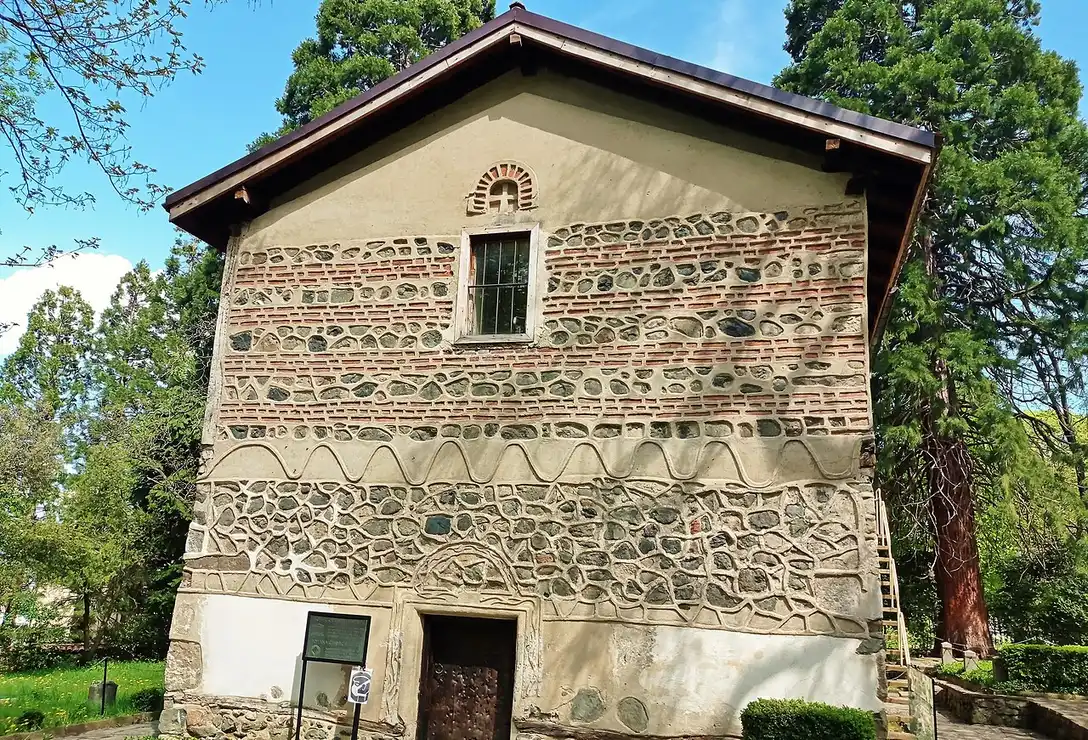 The stone and brick west facade of the small medieval Boyana Church, ringed by tall trees below Vitosha