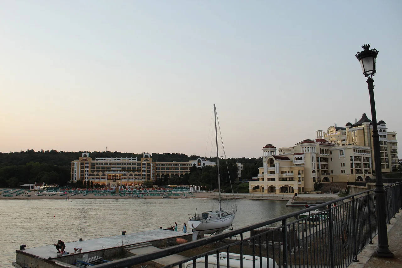 The hotels of Duni Royal Resort seen from the pier at dusk, a private beach lined with parasols below them and a yacht moored in the foreground