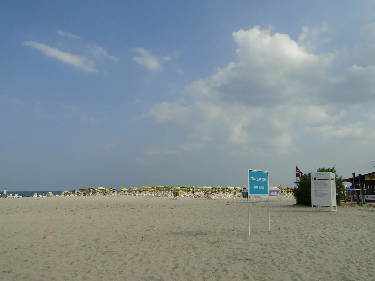A wide empty stretch of Bulgarian beach with a blue sign reading Free Zone and rows of paid yellow parasols in the distance