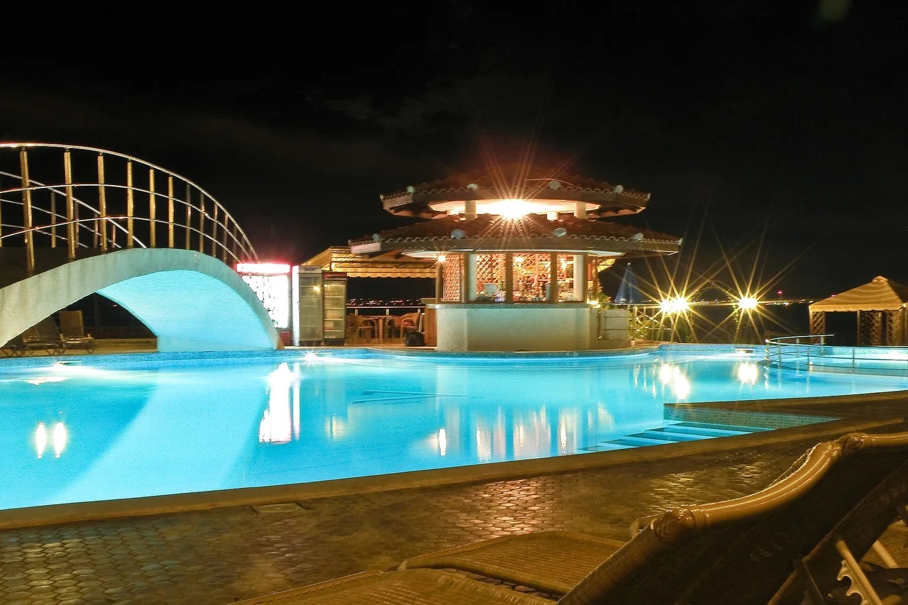 A floodlit hotel pool at night with a thatched pool bar behind it and empty sun loungers in the foreground