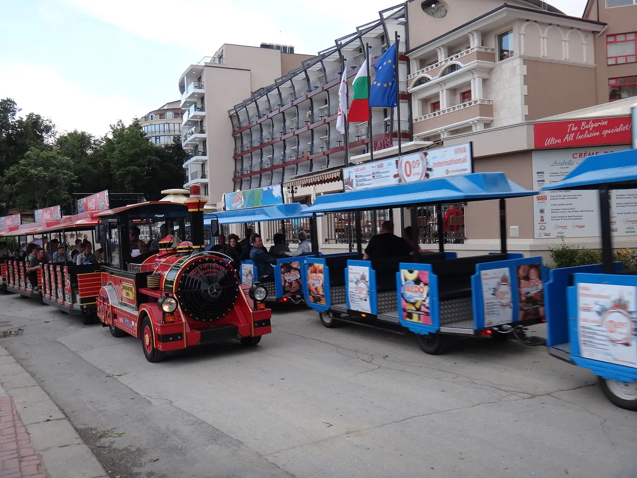 A hotel frontage at Golden Sands with Bulgarian and EU flags and a banner reading The Bulgarian Ultra All Inclusive specialist, a tourist land-train parked in front