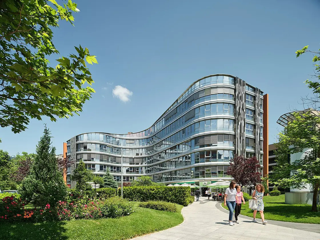 The curved glass facade of a modern office building at Business Park Sofia, with landscaped gardens and people walking past