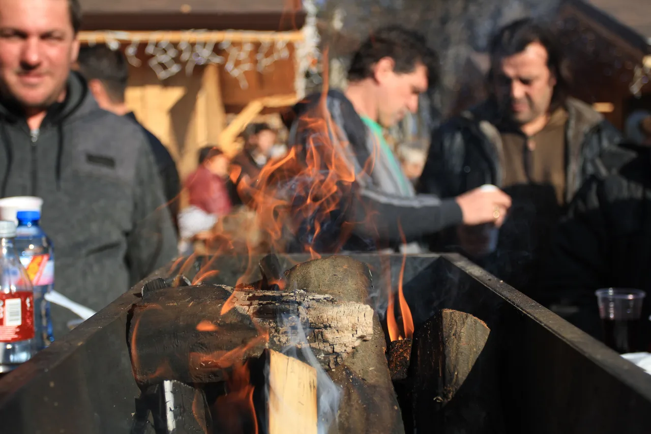 Logs burning in a long metal brazier at a Bulgarian Christmas market, with visitors warming their hands and wooden chalets behind