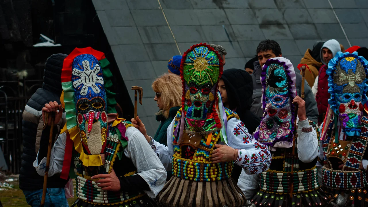 A line of kukeri in beaded and sequinned masks with rows of copper bells at their waists, waiting to parade at a Bulgarian masquerade festival