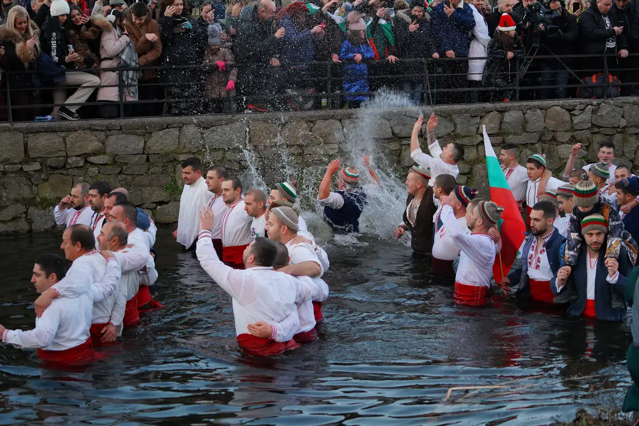 Men in white embroidered shirts standing waist-deep in the Tundzha river at Kalofer on Epiphany, arms locked, a Bulgarian flag raised and a crowd watching from the bank