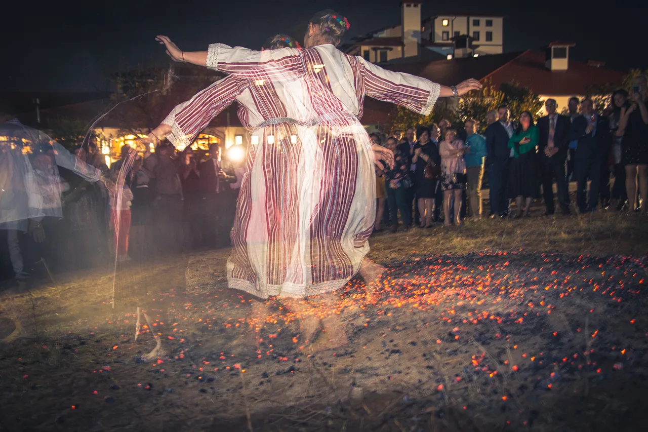 A dancer in striped Bulgarian folk dress stepping barefoot across a circle of glowing embers at night while a crowd watches from the edge