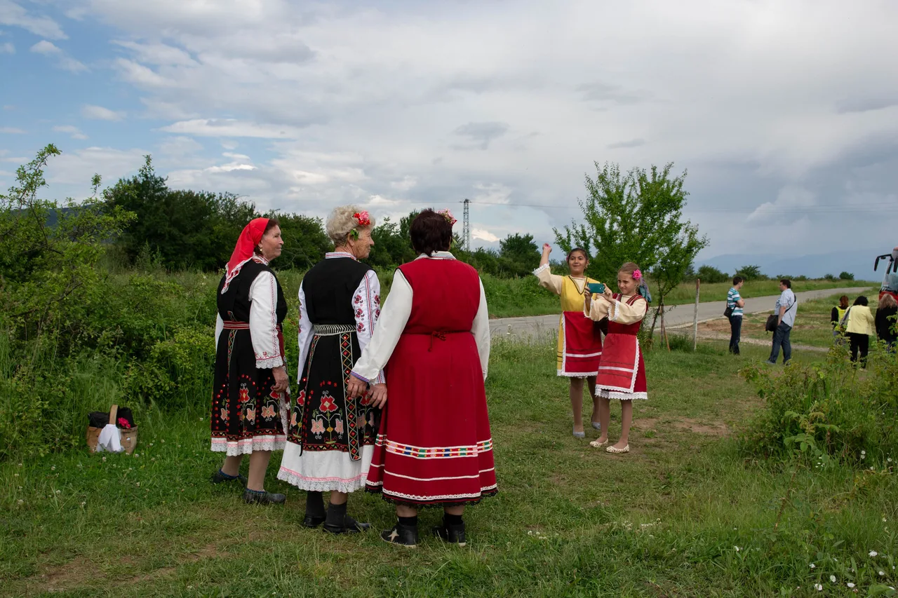 Three older women in embroidered Bulgarian folk dress standing at the edge of a rose field near Kazanlak while two girls in costume photograph them