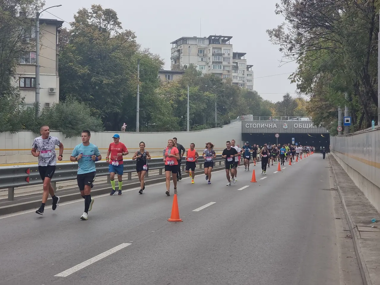 A field of runners in race bibs coming out of a road tunnel in central Sofia during the Sofia Marathon, traffic cones dividing the carriageway