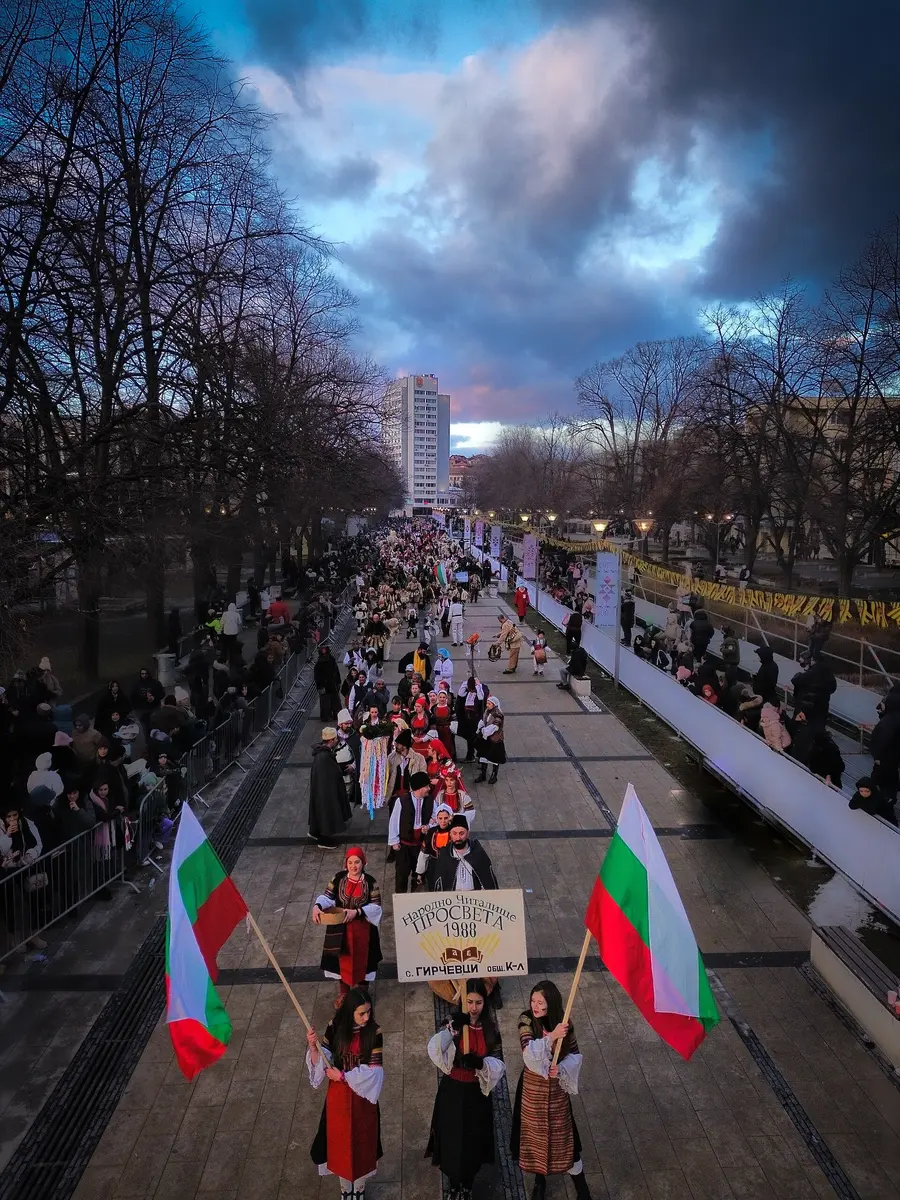A long parade column in folk costume moving down a barricaded pedestrian avenue in Pernik at dusk, two Bulgarian tricolour flags carried at the front