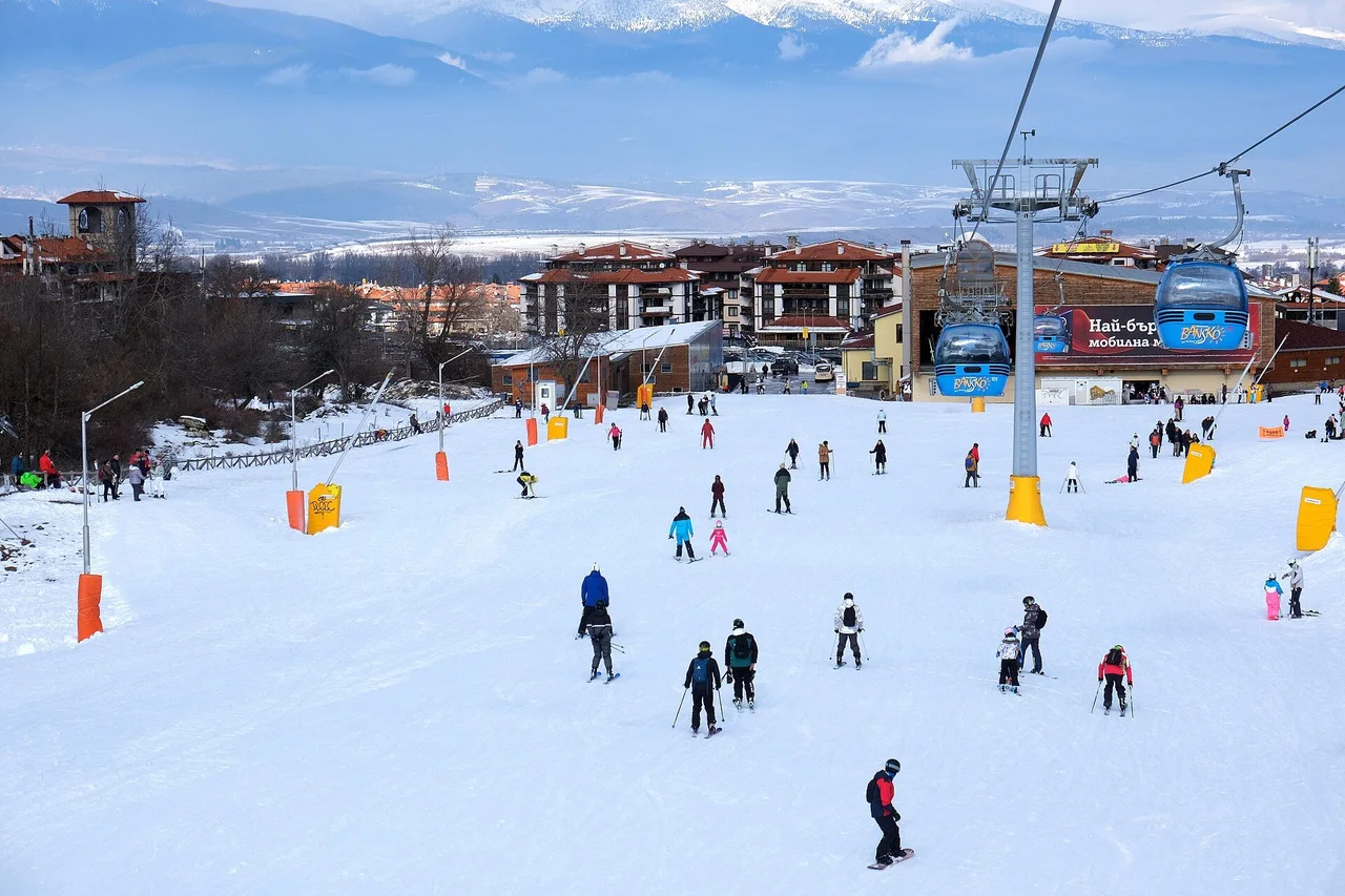 Skiers and snowboarders on the wide nursery slope at the foot of Bansko, with two gondola cabins passing above the base station and the town and valley behind