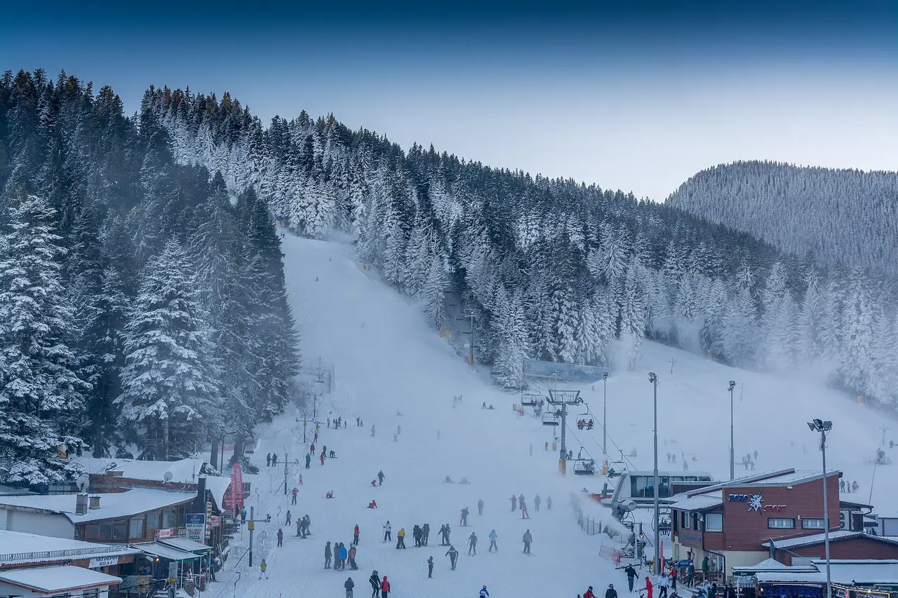The floodlit base area at Borovets at dusk, with skiers on the piste running right up to the hotels and shops and a snow cannon working on the right