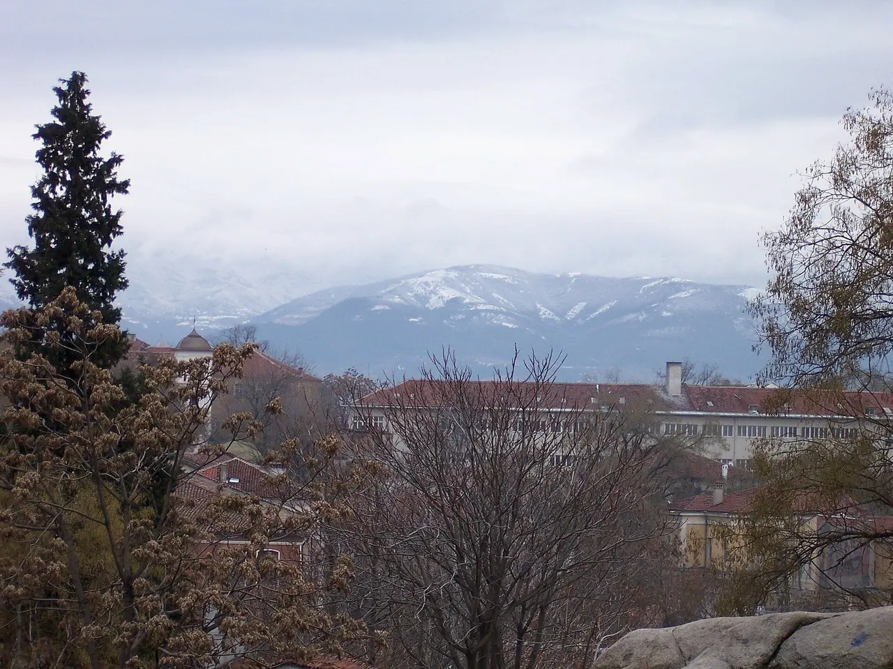 Red-tiled roofs of Plovdiv under a heavy grey sky with the snow-dusted Rhodope foothills rising behind the city