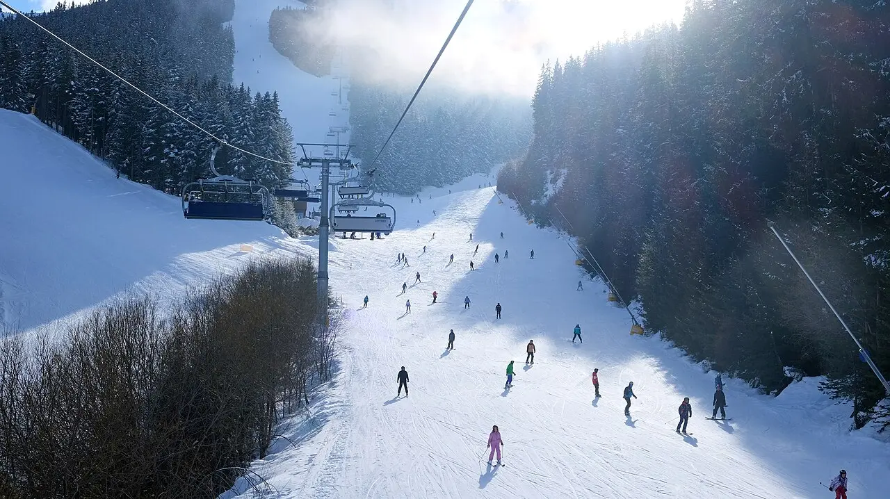 A chairlift climbing a tree-lined piste at Bansko with skiers and snowboarders descending through sunlit snow below
