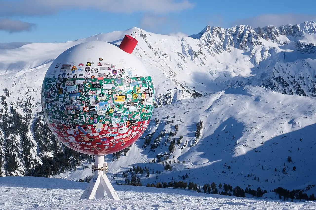 A large sticker-covered globe landmark at a high point in Bansko, overlooking open, ungroomed mountainside beyond the marked pistes