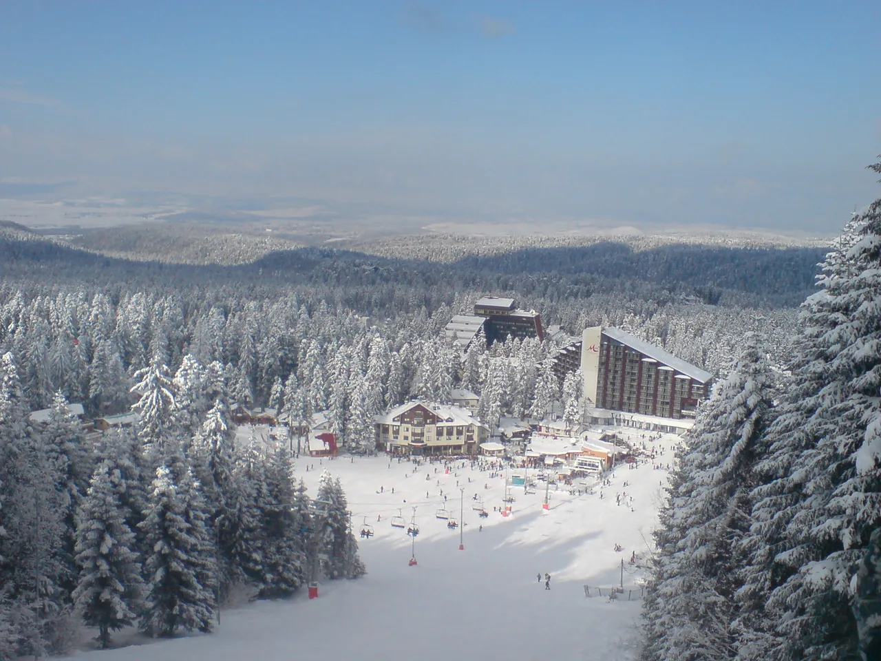 Borovets in winter seen from the lower slopes: the hotel base at the foot of the pistes, snow-covered pine forest and the Samokov plain in the distance