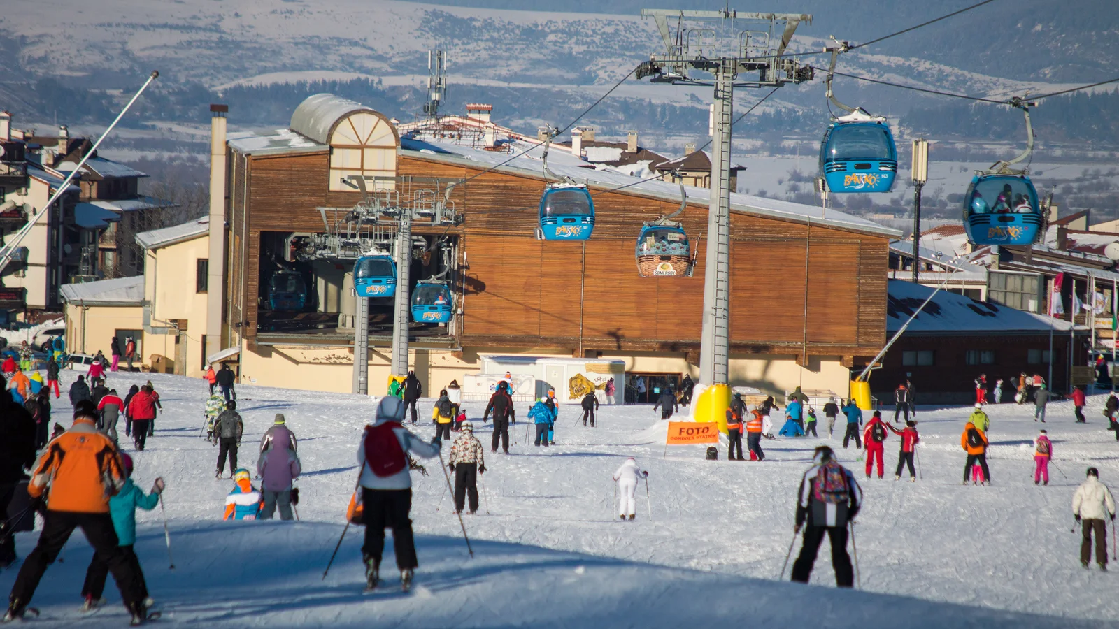 A blue gondola cabin marked Bansko arriving at a wooden lift station above a busy ski slope, skiers gathered below and snow-covered peaks behind