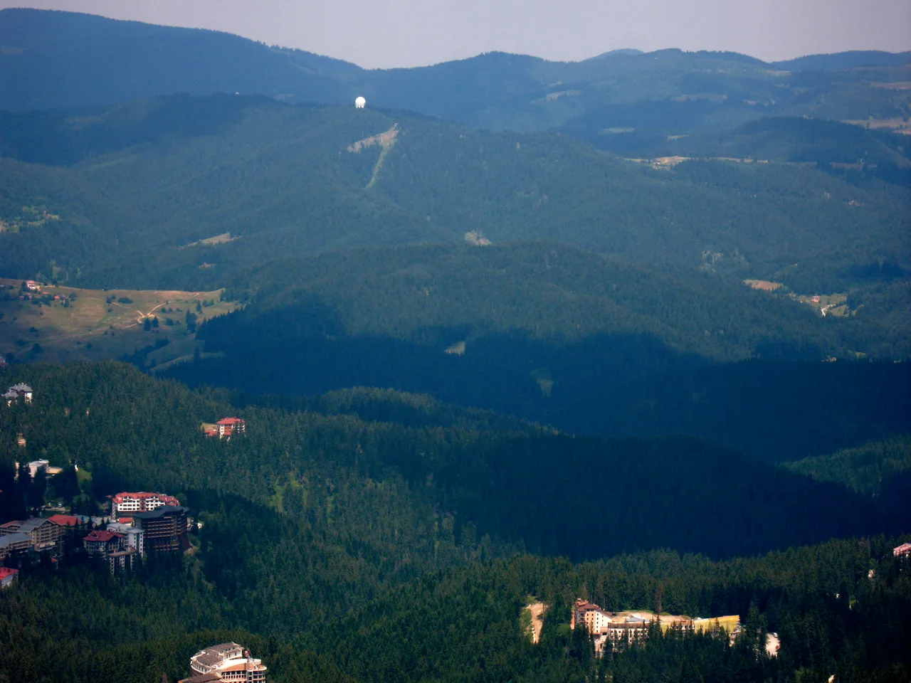 Pamporovo hotels among the pine forest seen from Snezhanka peak, the rounded Rhodope ridges and a white observatory dome behind