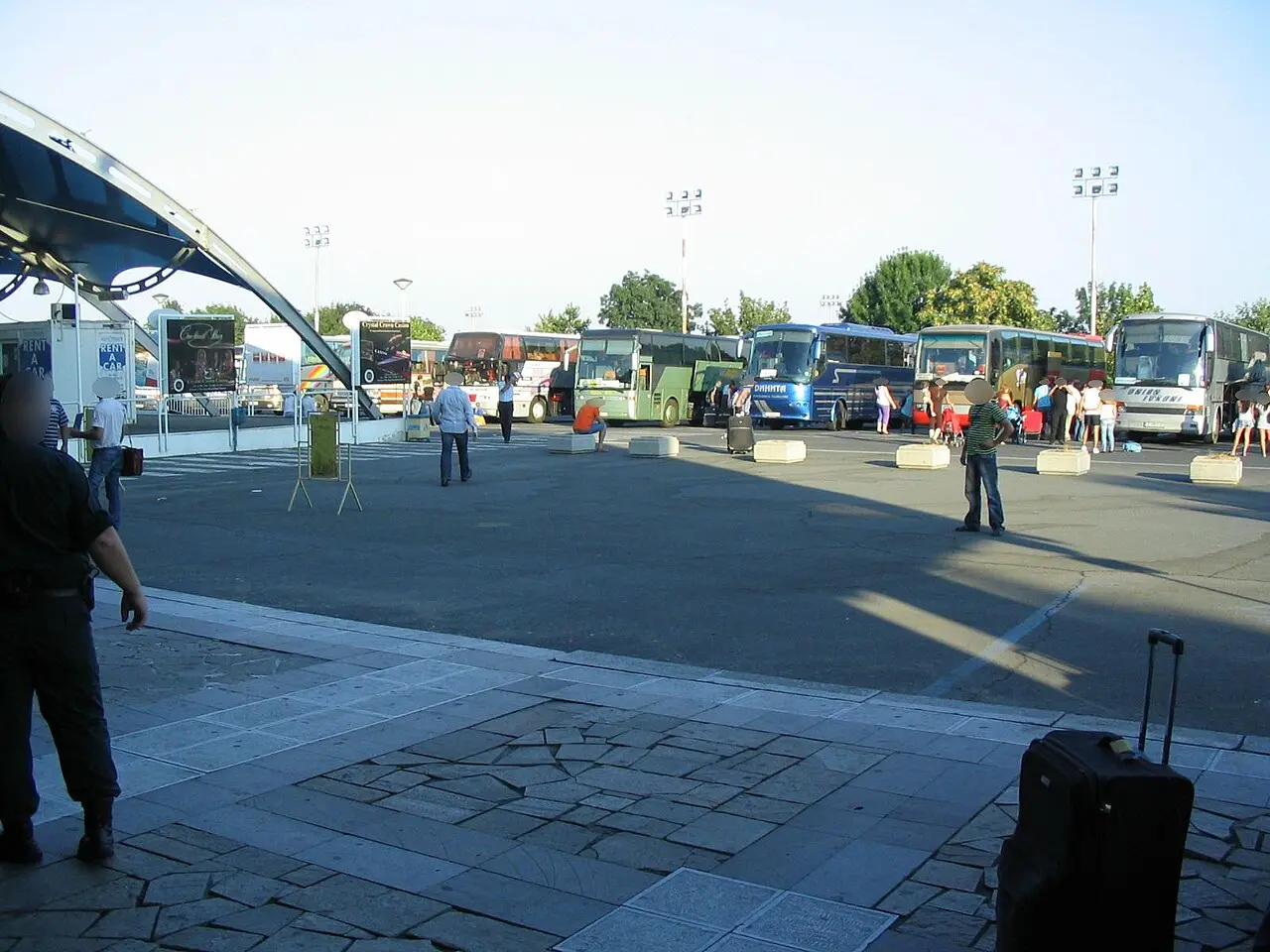 A row of tour coaches and a rent-a-car booth in the coach park just outside the Burgas Airport exit, with arriving passengers and luggage