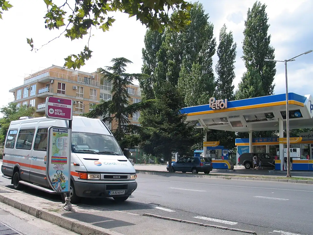 A Sunny Beach bus stop sign and a resort shuttle minibus parked on a street beside a petrol station