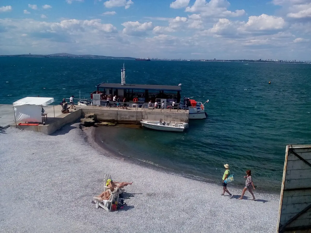A pebbly cove and jetty on St Anastasia Island with the excursion boat moored, and the skyline and port of Burgas across the bay on the horizon
