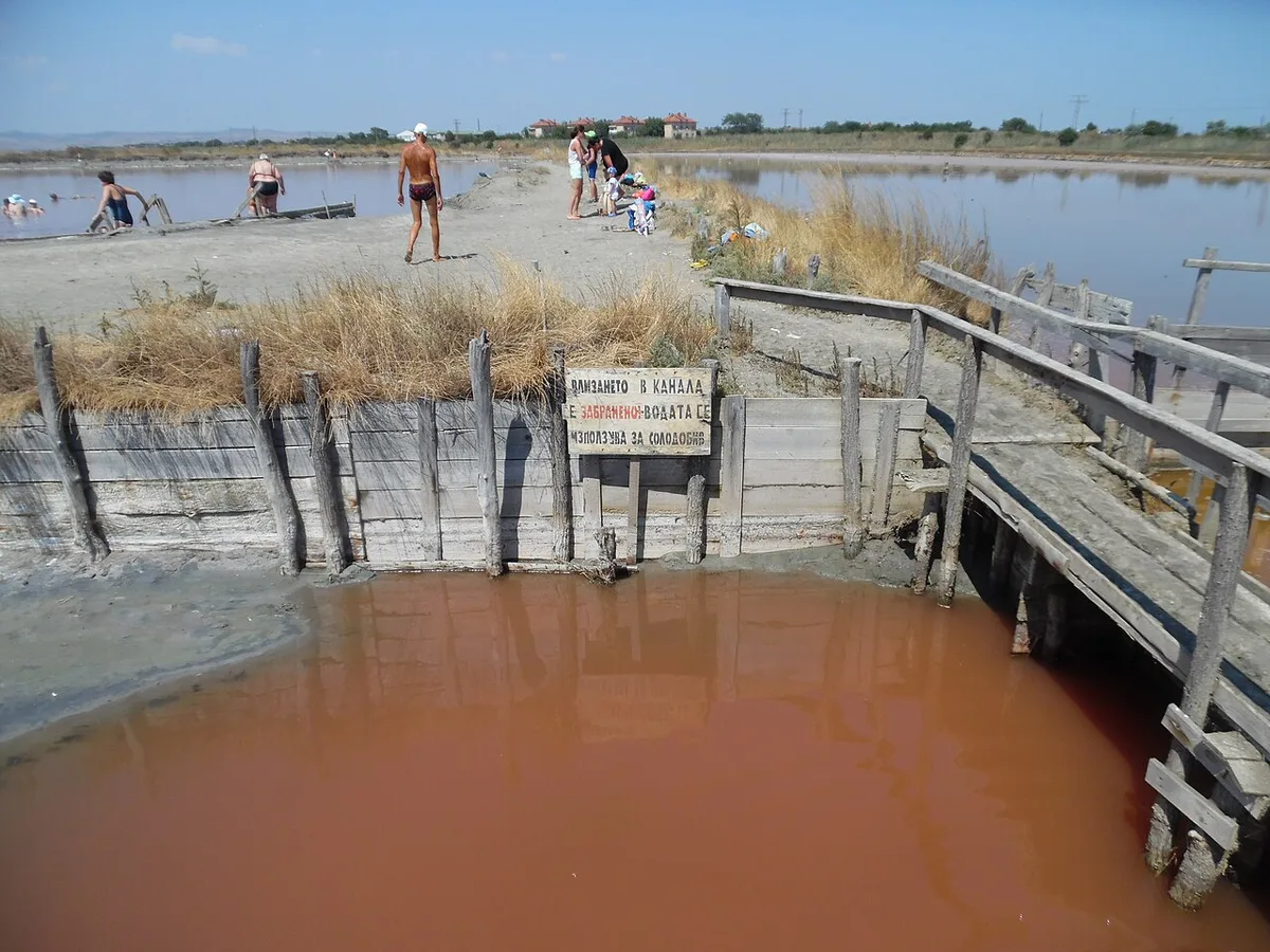 A channel of rusty-pink saline water at the Atanasovsko salt lake near Burgas, with wooden walkways, and people on the sandy dyke behind bathing in the salt mud