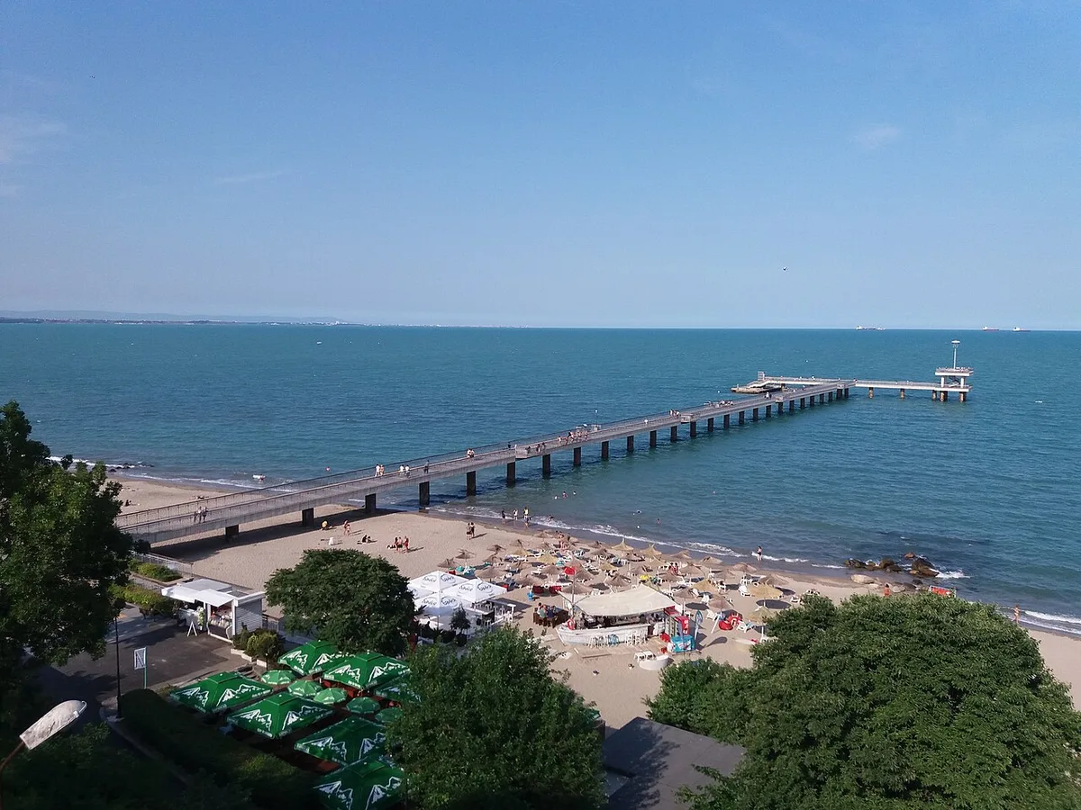The long Burgas sea bridge reaching out into the blue Black Sea from the Central Beach, with sun umbrellas and beach bars on the sand below