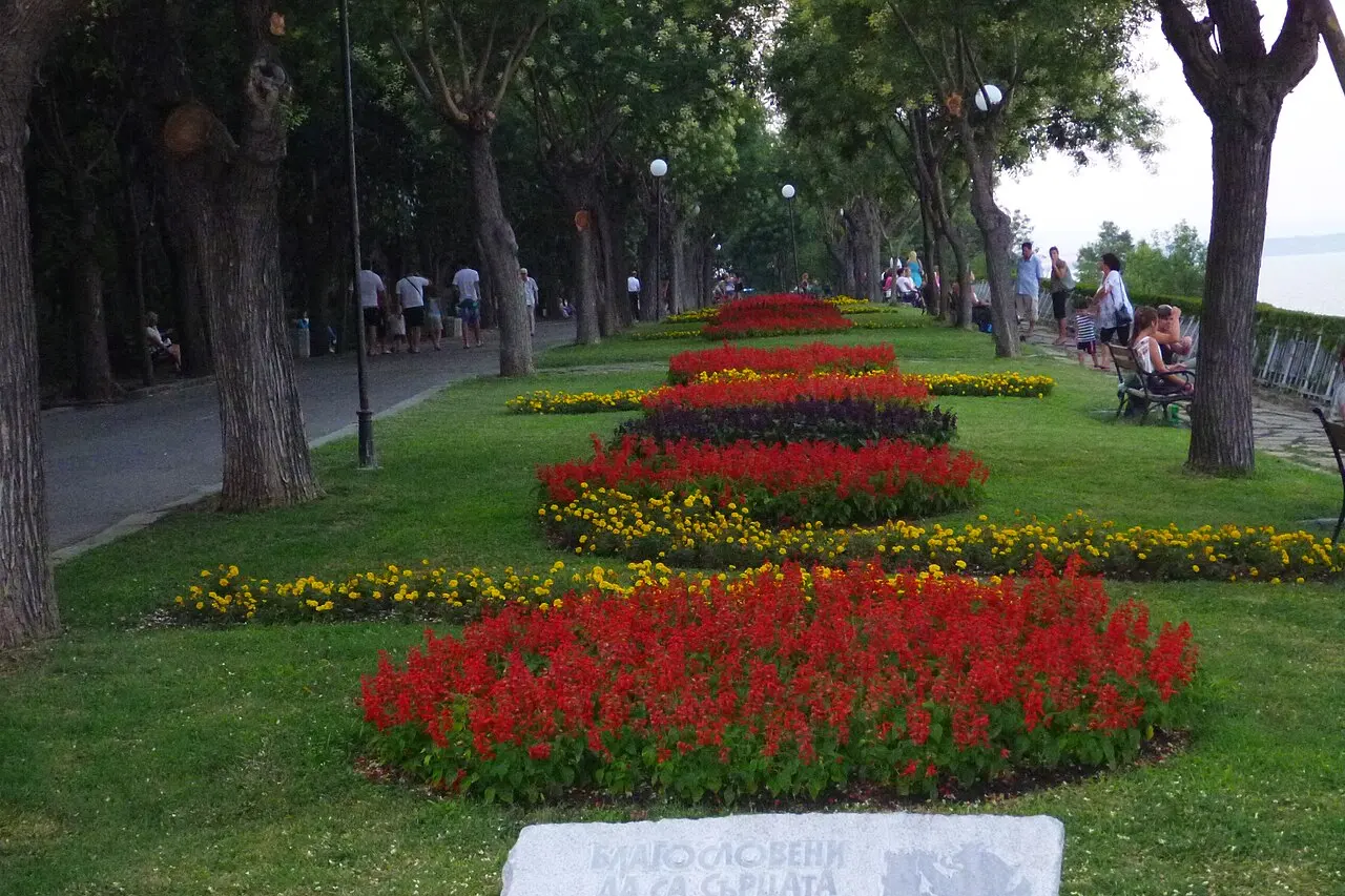 A long flowerbed of red and yellow blooms running down a tree-lined alley in the Sea Garden of Burgas, with people walking and the sea to one side