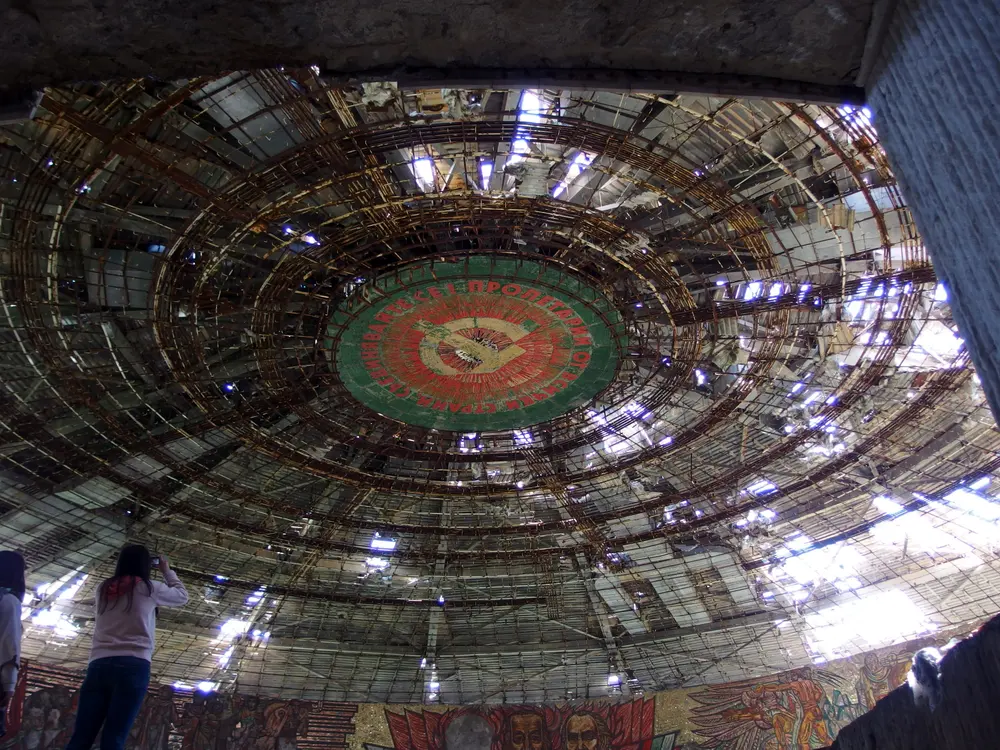 The circular hall inside the Buzludzha monument seen from below, its ceiling ring of mosaic and stripped roof panels open to daylight