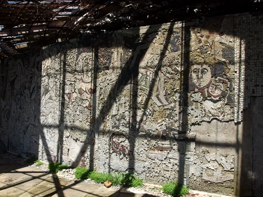 A ruined mosaic wall inside the Buzludzha monument with faces of figures still visible above fallen tesserae and weeds