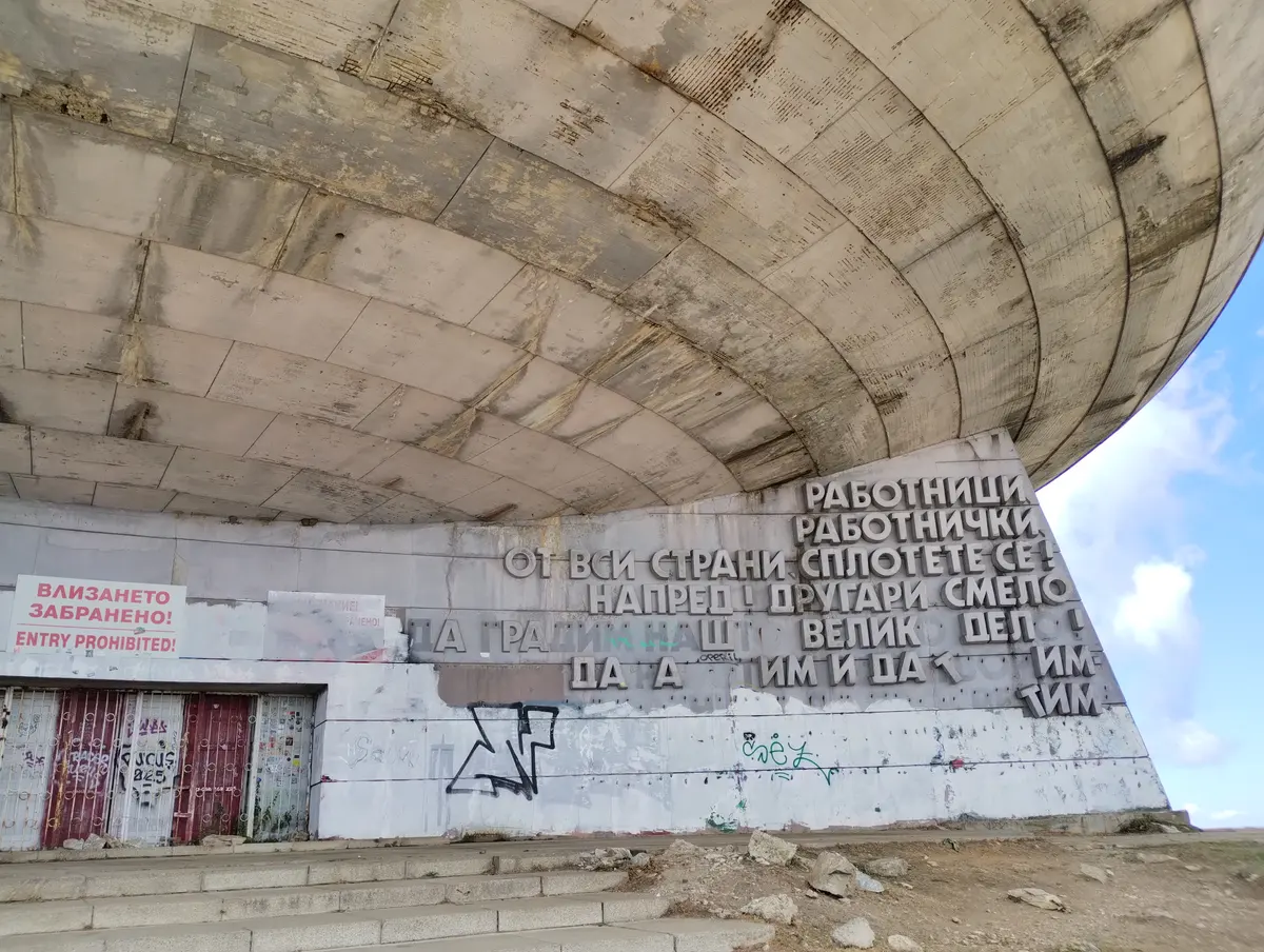 The sealed lower entrance of the Buzludzha monument beneath the overhanging concrete saucer, with red-painted doors, graffiti and faded slogans