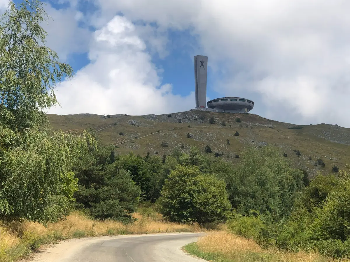 A narrow mountain road curving up a grassy ridge with the Buzludzha monument silhouetted on the skyline ahead